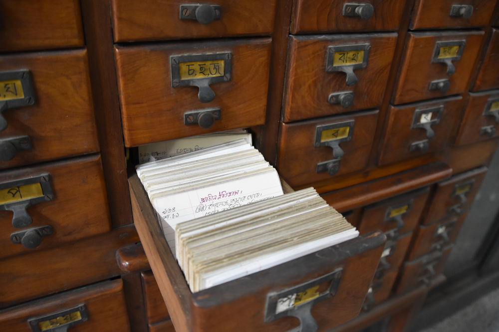 A wooden library card catalog with one drawer pulled open, revealing a stack of index cards written in Bengali script.