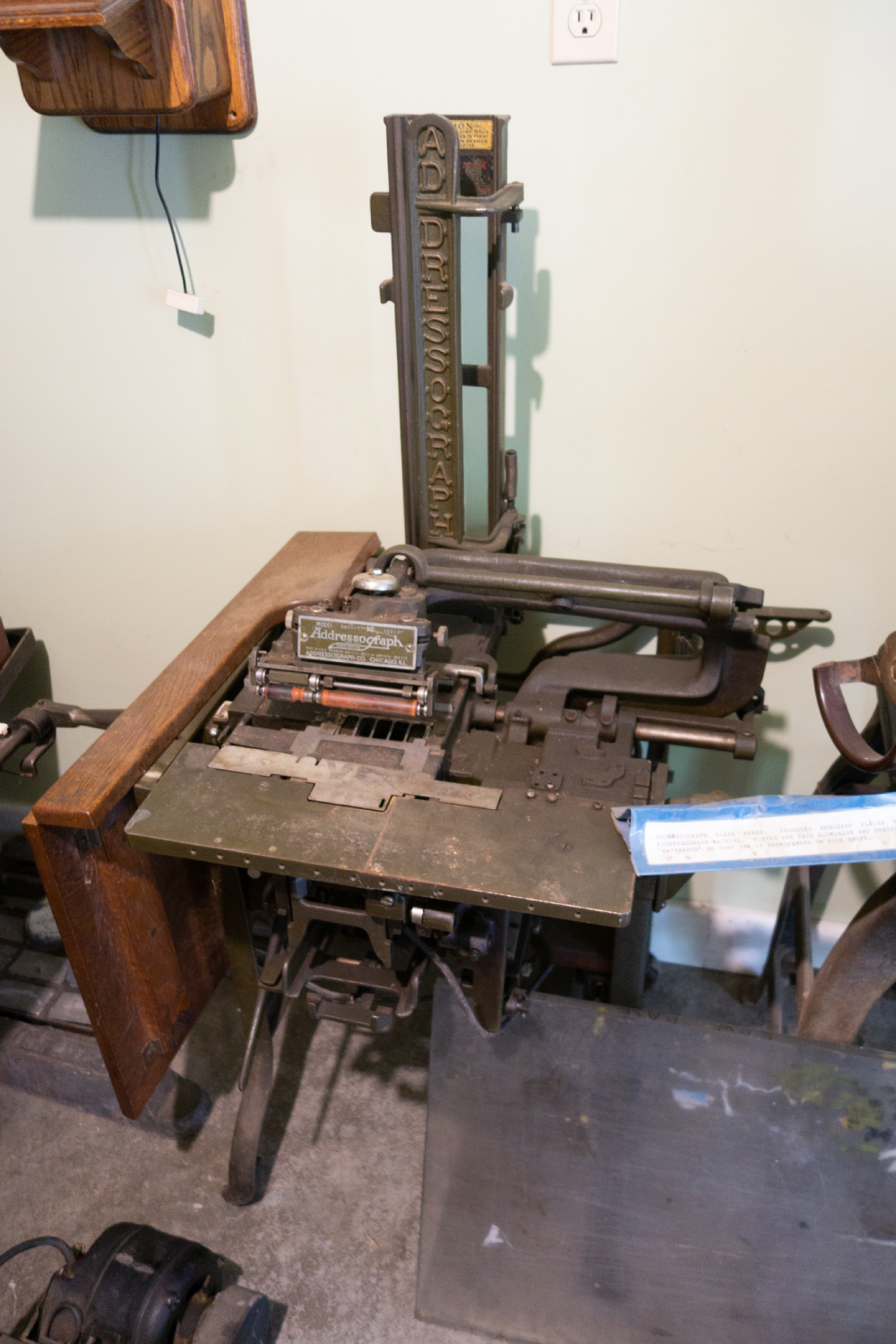 Vintage Addressograph machine with a vertical metal plate and wooden side table.