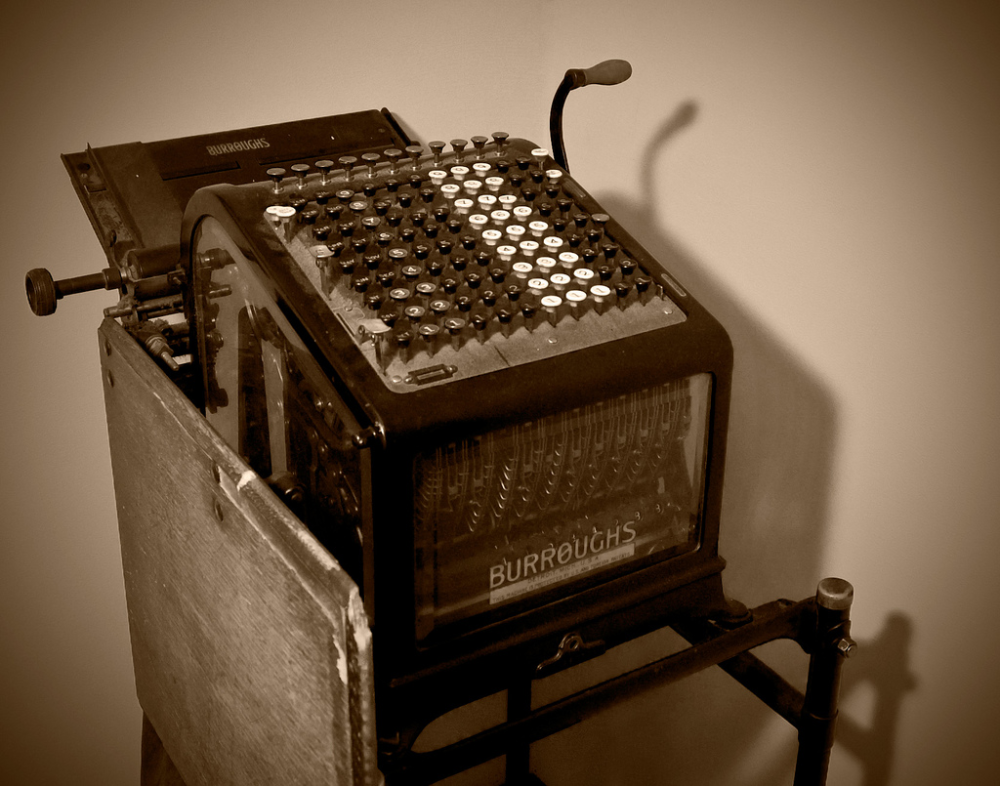Vintage Burroughs adding machine with a manual lever and numerical keypad on a metal stand.