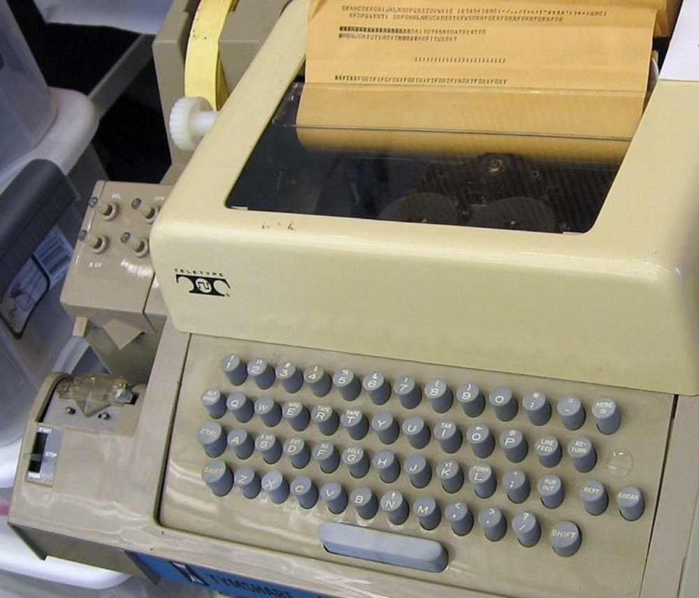 A beige Teletype machine with a QWERTY keyboard and a paper roll showing printed text.