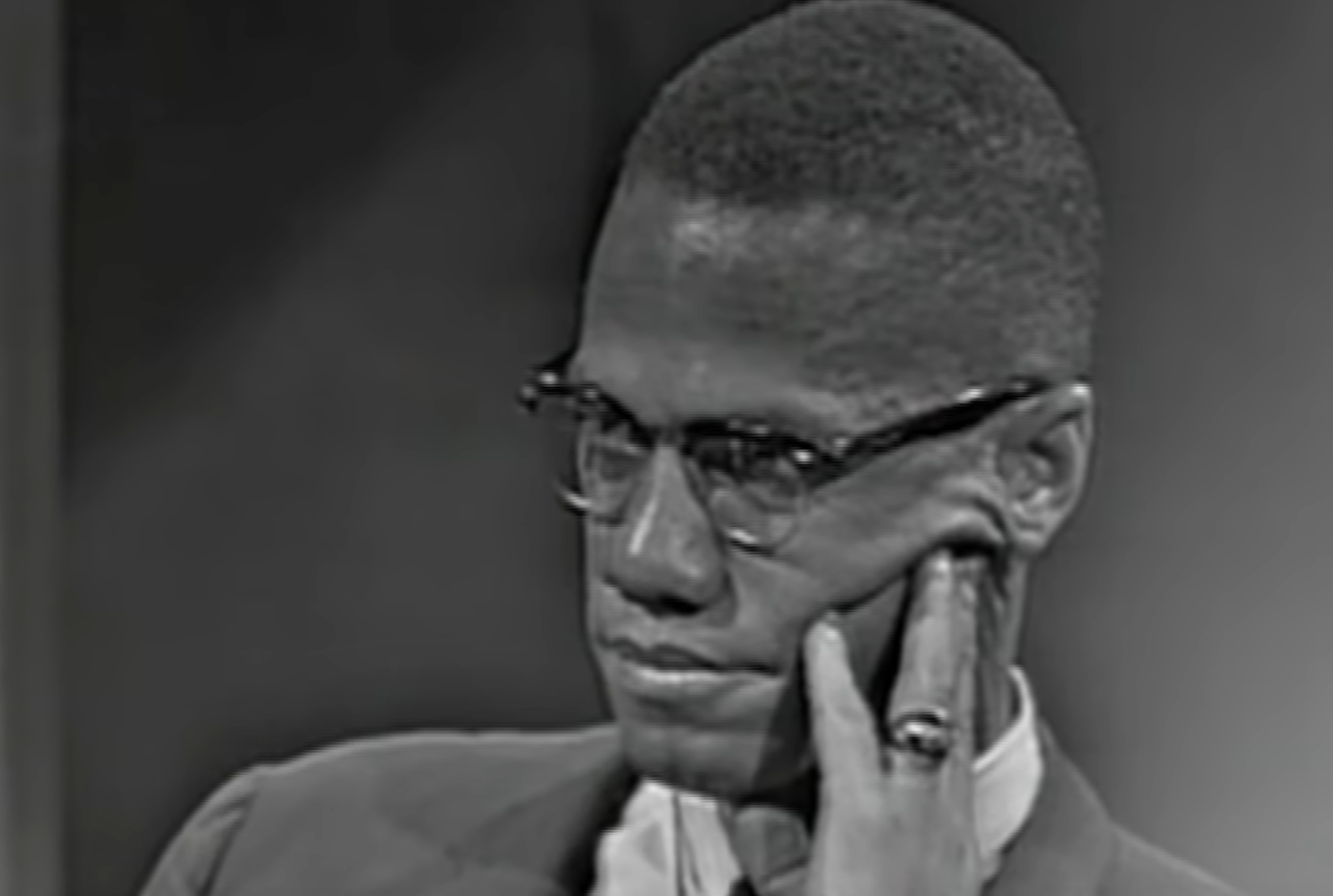 Malcolm X wearing glasses and a suit, resting his hand against his cheek.