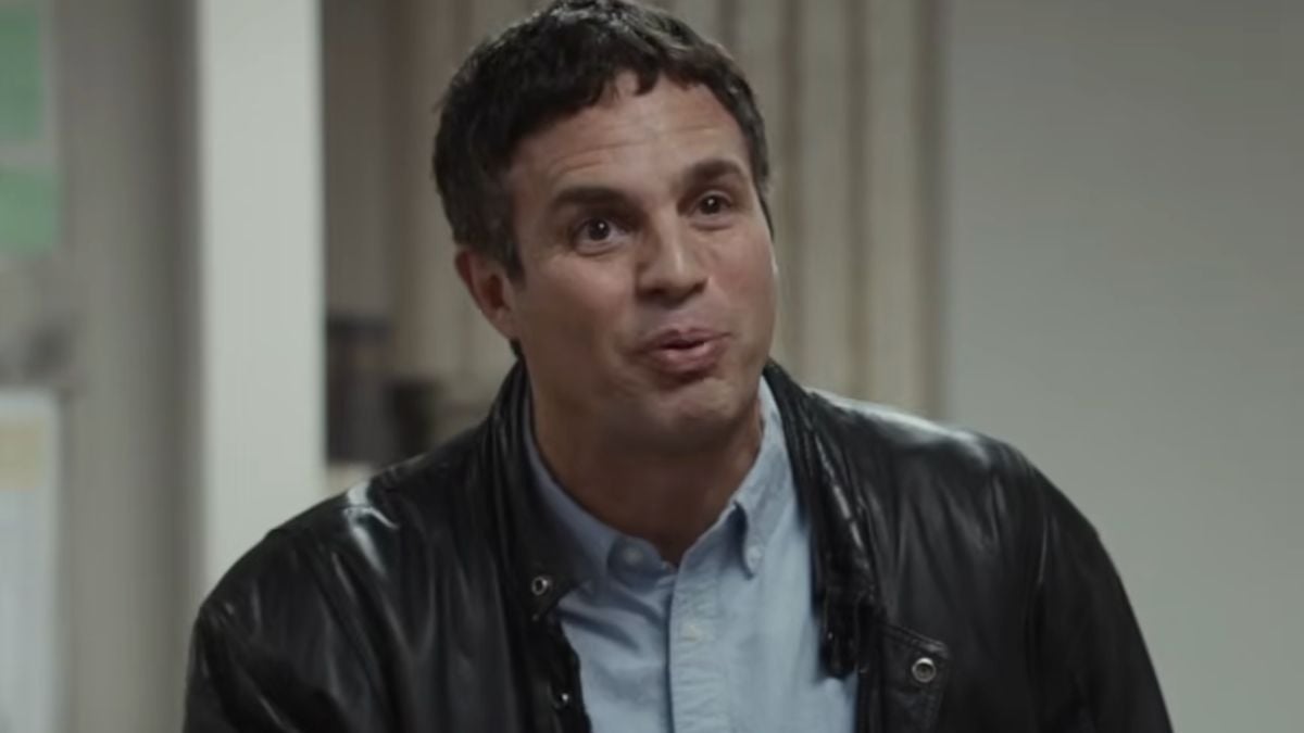 A man with short dark hair wearing a black leather jacket over a light blue shirt is indoors, looking slightly upward with a neutral expression.