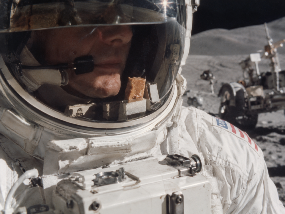 An astronaut seen through a helmet visor, with a piece of food attached to the inside of the helmet near their mouth, and a lunar rover in the background.