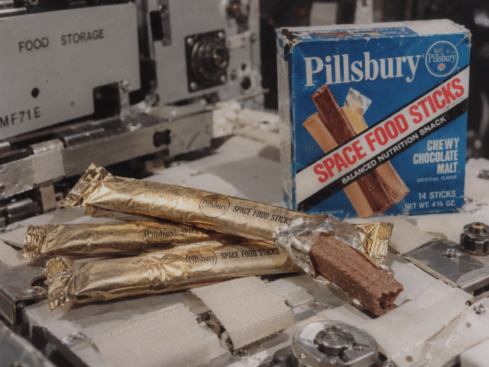 A box of Pillsbury Space Food Sticks and several individually wrapped sticks resting on a piece of space station equipment.