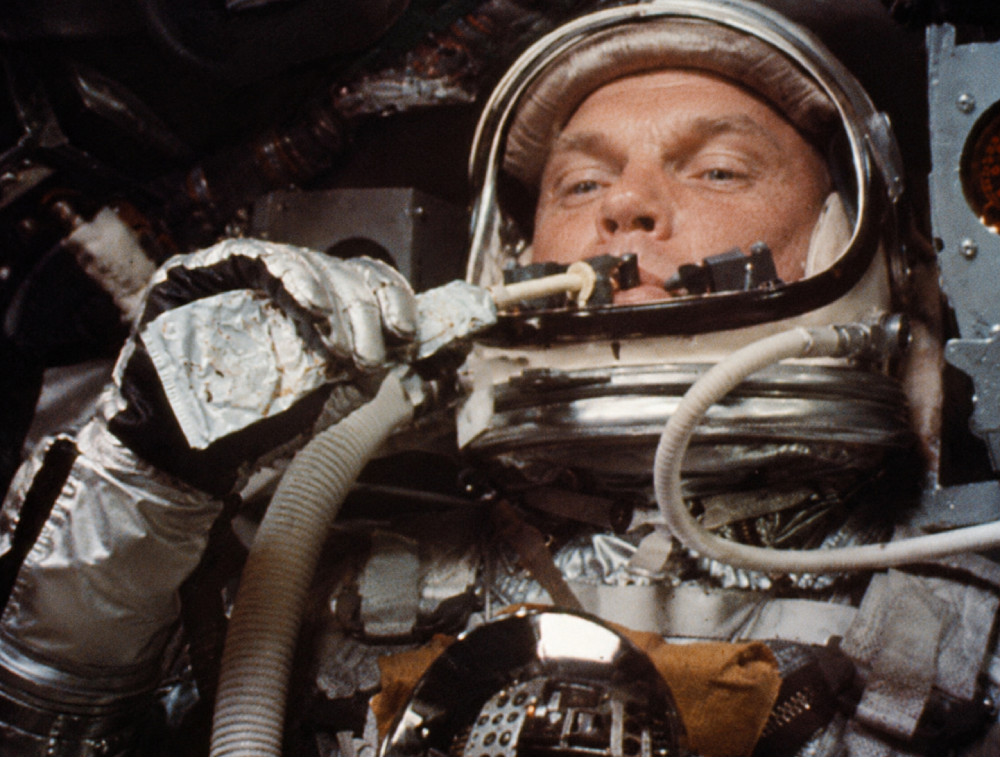 An astronaut in a spacesuit helmet eats food from a tube.
