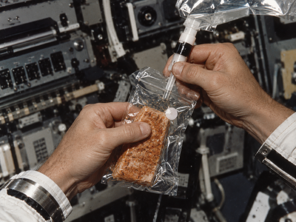 Astronaut hands injecting water into a sealed pouch containing a piece of space food.