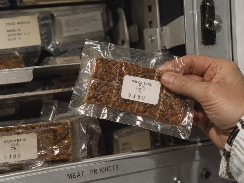 A hand holds a vacuum-sealed package of bacon bars in front of a metal storage unit labeled “Meat Products” and “Food Module.”