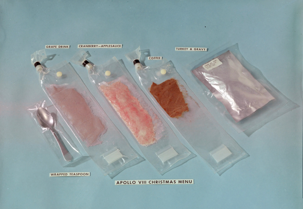 Apollo VIII space food rations in plastic pouches labeled Grape Drink, Cranberry-Applesauce, Coffee, and Turkey & Gravy, with a wrapped teaspoon.
