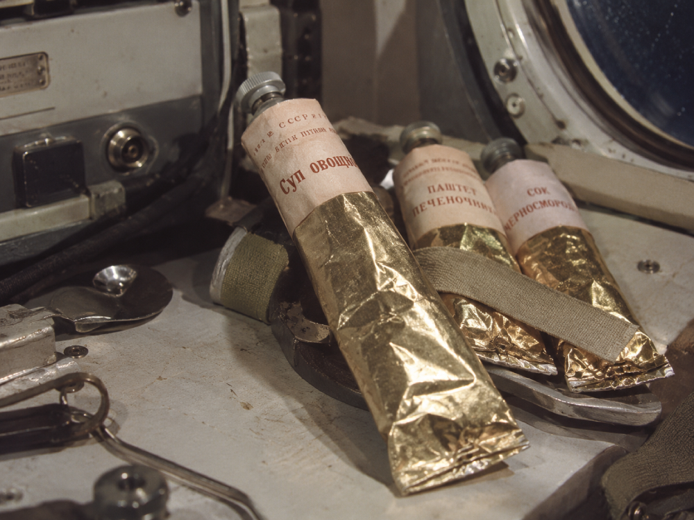 Three gold-wrapped food tubes with Russian text labels resting on a spacecraft instrument panel.