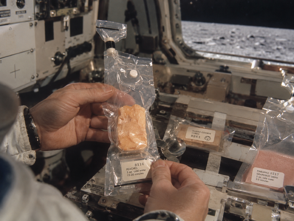 An astronaut holds a plastic pouch of freeze-dried peaches inside a spacecraft, with the lunar surface visible through the window.