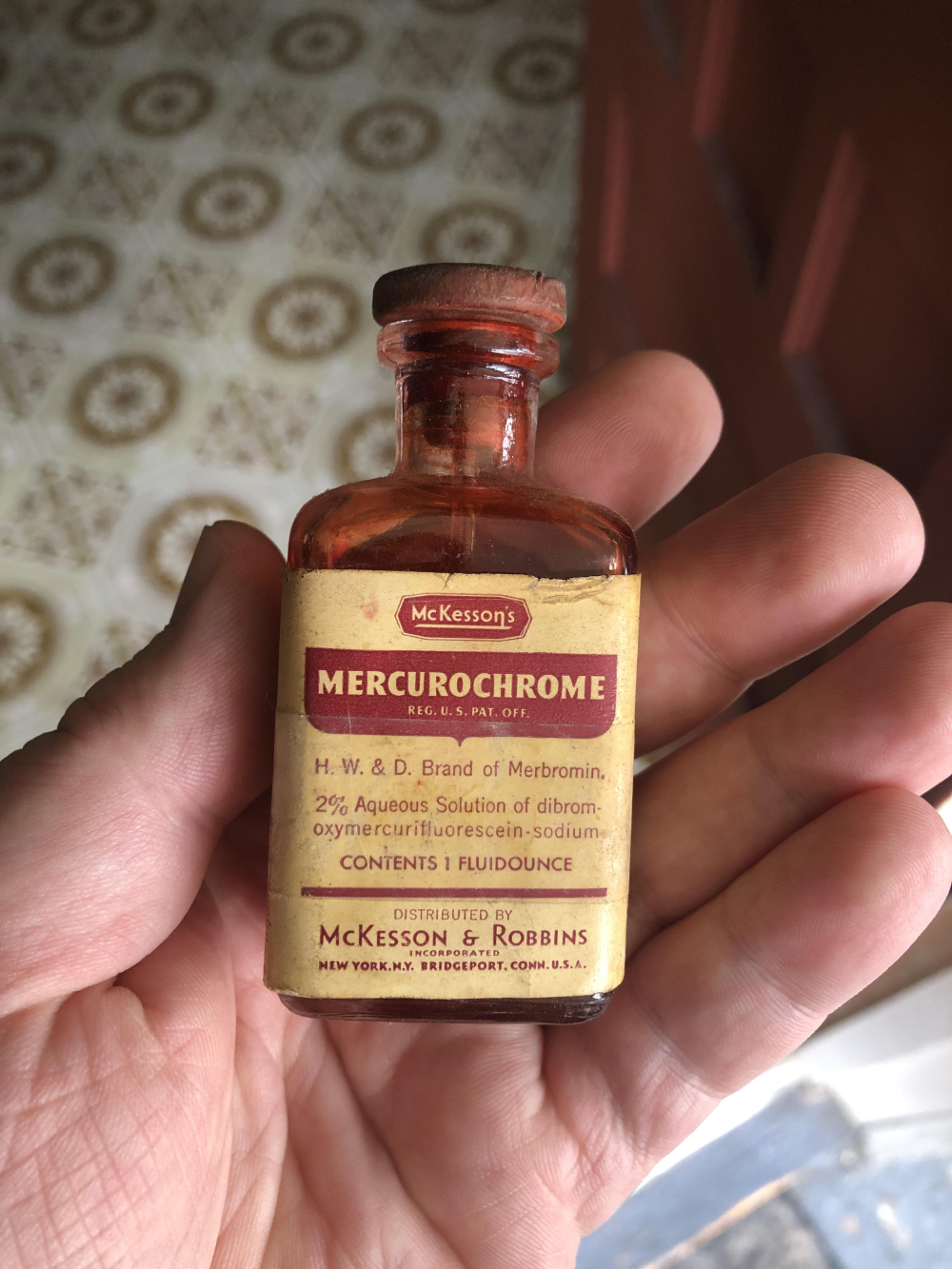 A hand holding a small, amber glass bottle of McKesson’s Mercurochrome with a cork stopper and a paper label listing the ingredients and manufacturer.