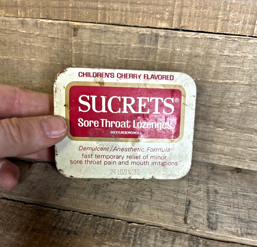 A hand holding a tin of Children’s Cherry Flavored Sucrets sore throat lozenges.