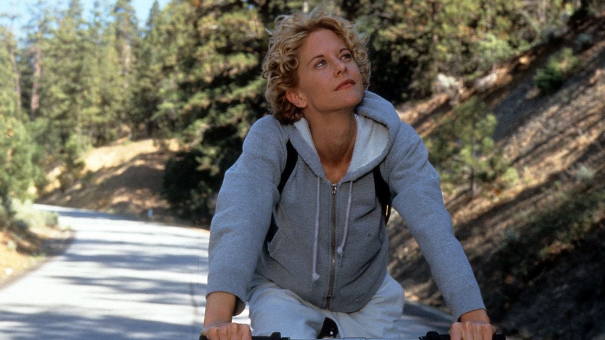 A woman with short blonde hair, wearing a gray hoodie and white pants, rides a bicycle on a road through a forested area, looking up thoughtfully.