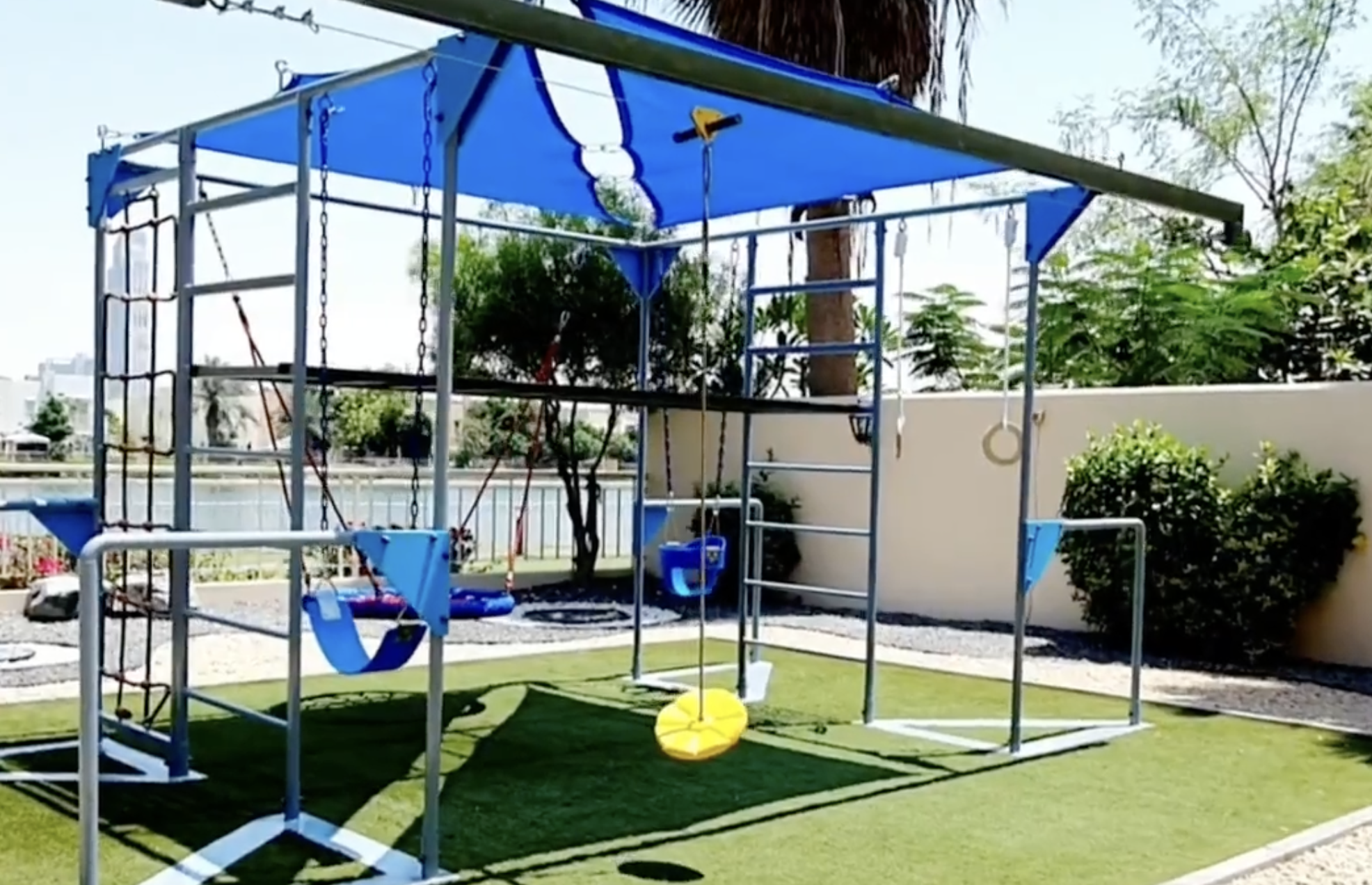A backyard metal play gym with a blue canopy, including a swing, a tire swing, gymnastic rings, a rope climber, and a ladder, situated on a patch of artificial turf.