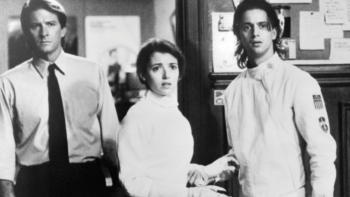 Three people stand indoors looking concerned; one man wears a dress shirt and tie, a woman and another man wear fencing uniforms. The background features a bulletin board and blurred posters.