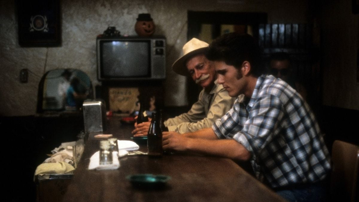 Two men sit at a dimly lit bar counter; one older with a mustache and hat, the other younger in a plaid shirt, both looking down. A TV and Halloween pumpkin decoration are in the background.