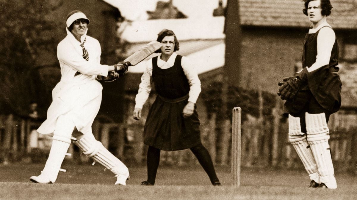 Three women play cricket outdoors in vintage attire; one bats, one stands behind with gloves and pads, and one is a wicketkeeper. The scene appears historical, with old-fashioned uniforms and buildings in the background.