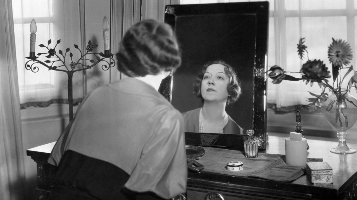 A woman with short, wavy hair sits at a vanity table, looking at her reflection in a mirror. The table holds various items, and sunlight filters through the window behind her.