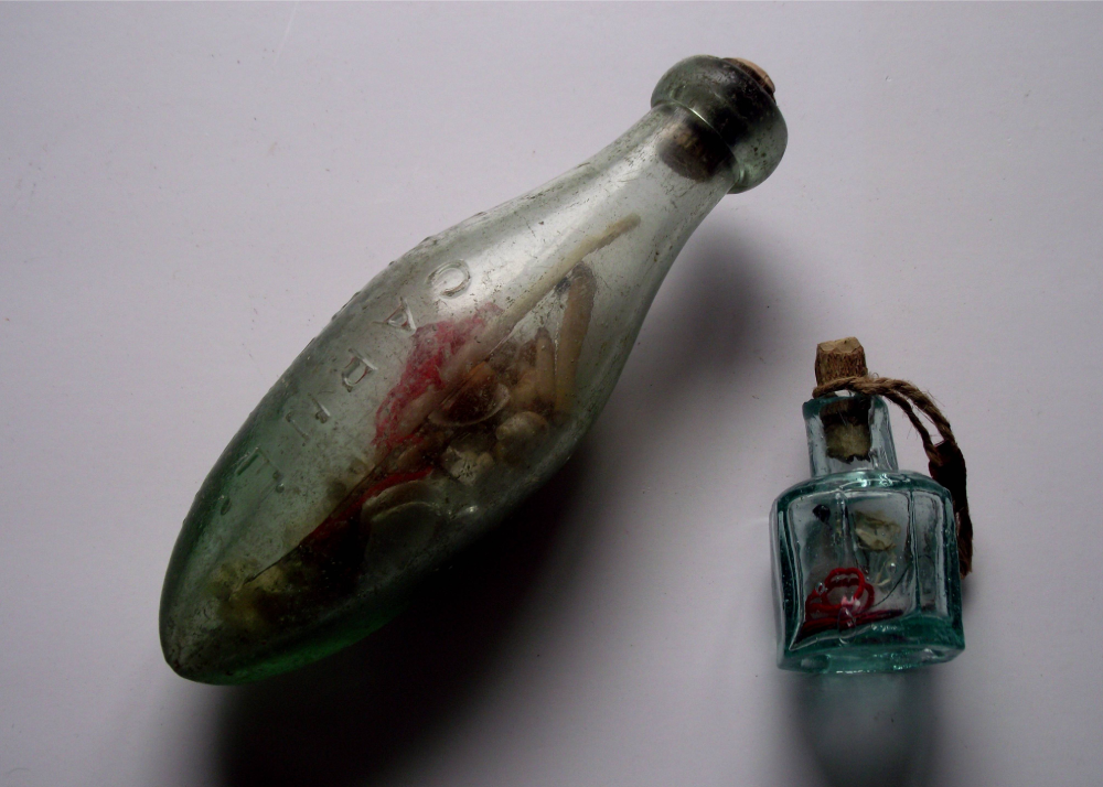A large, oblong glass bottle with a cork stopper and embossed text containing seashells, next to a smaller, square glass bottle with a cork and twine containing small items and a red object.