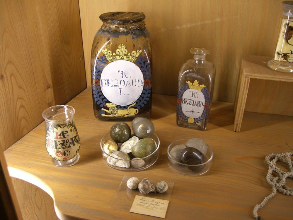 Glass jars and containers labeled “Bezoard” displayed on a wooden shelf, alongside various stone-like specimens and a handwritten label.