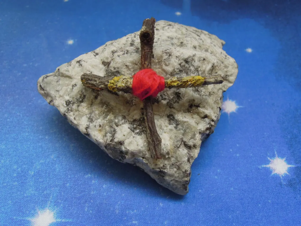 A cross made of two small, lichen-covered twigs bound together with red yarn, resting on a white speckled rock against a blue background with white stars.
