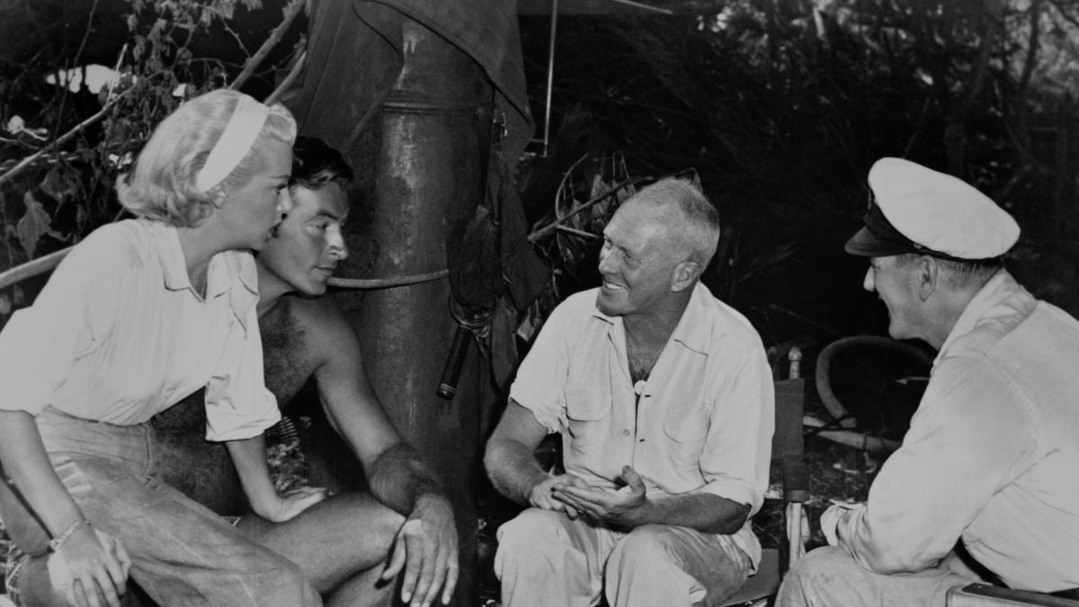 A woman and three men sit outdoors in a wooded area, engaged in conversation.