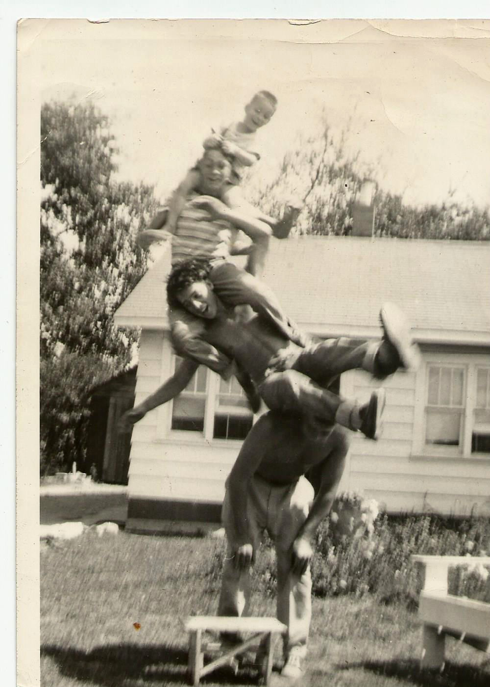 Three people stacked vertically in a human pyramid in a yard.