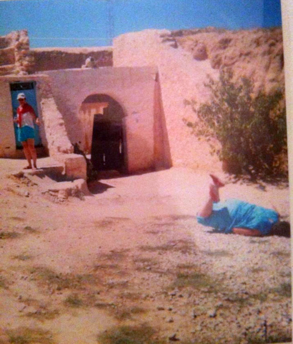 A person stands in the doorway of a tan desert structure while another person lies on the ground nearby.