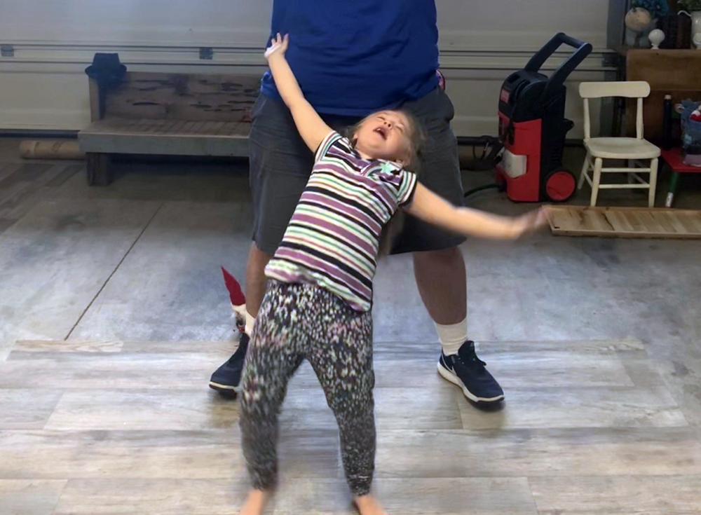 A young girl in a striped shirt and patterned leggings leans back with her arms outstretched while standing in front of an adult.
