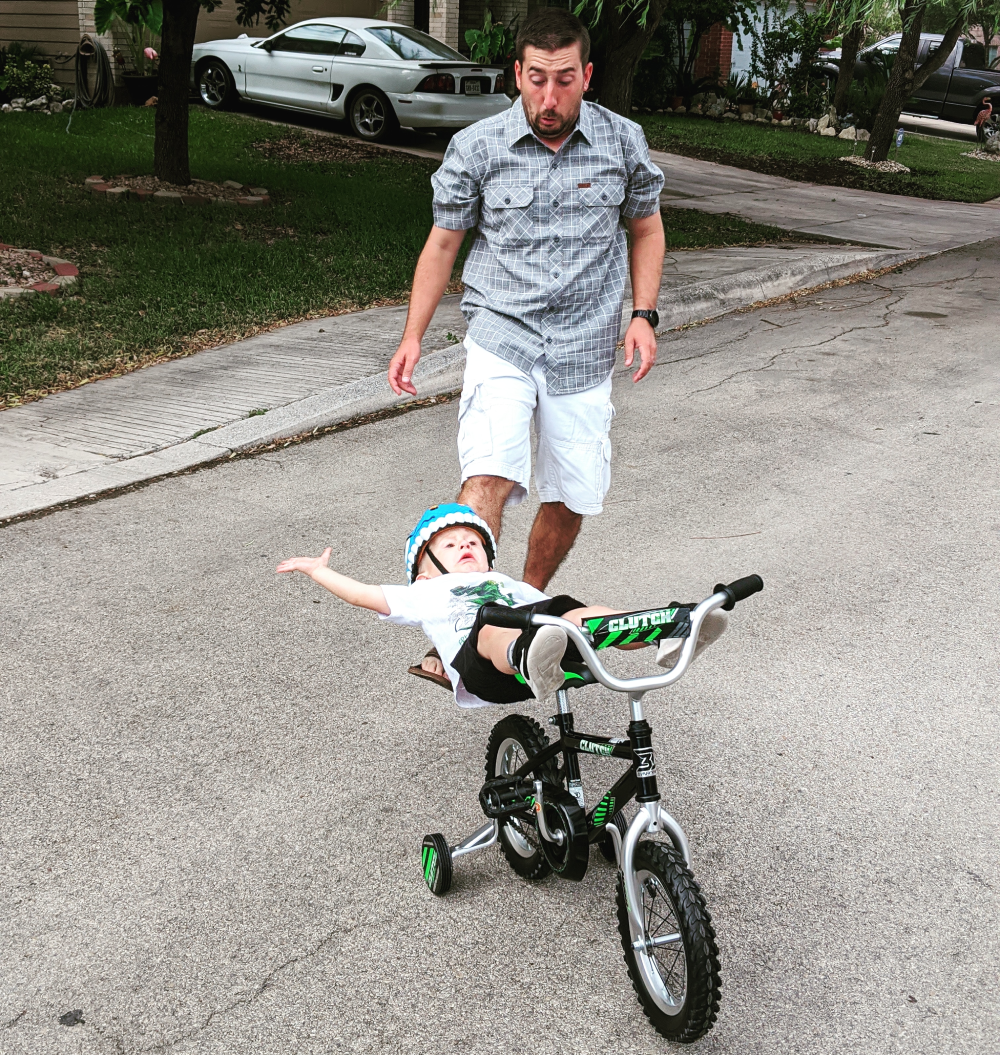 A man in a plaid shirt watches as a toddler in a bicycle helmet rides a small bike with training wheels while lying flat on their back.
