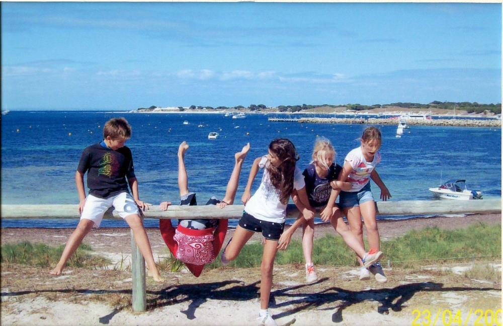 Four children balance on a wooden fence overlooking a bay, with one child hanging upside down from the rail.