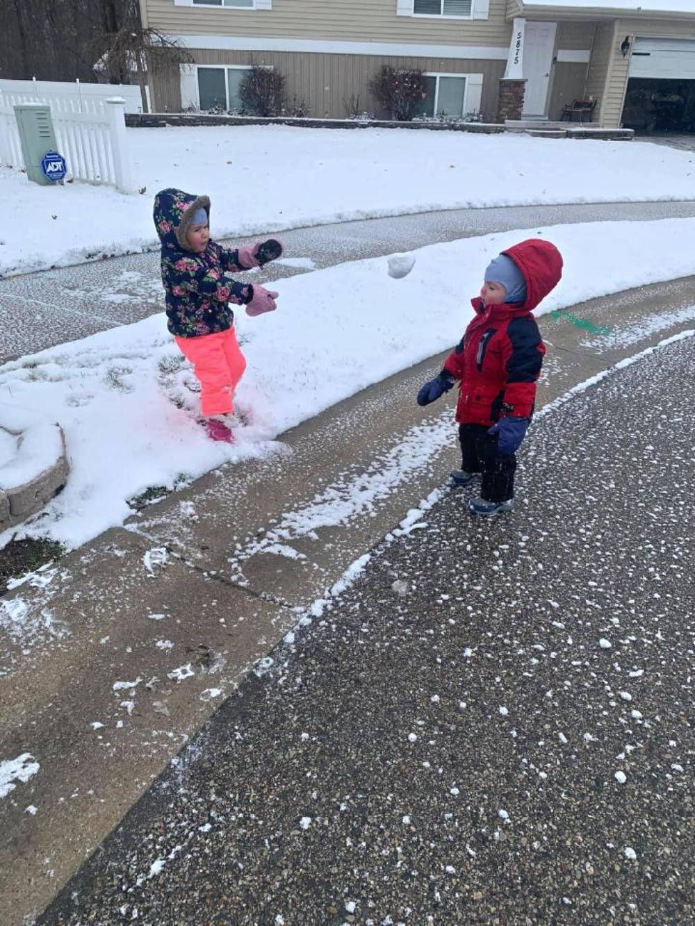 Two children in winter gear have a snowball fight on a snowy suburban street.
