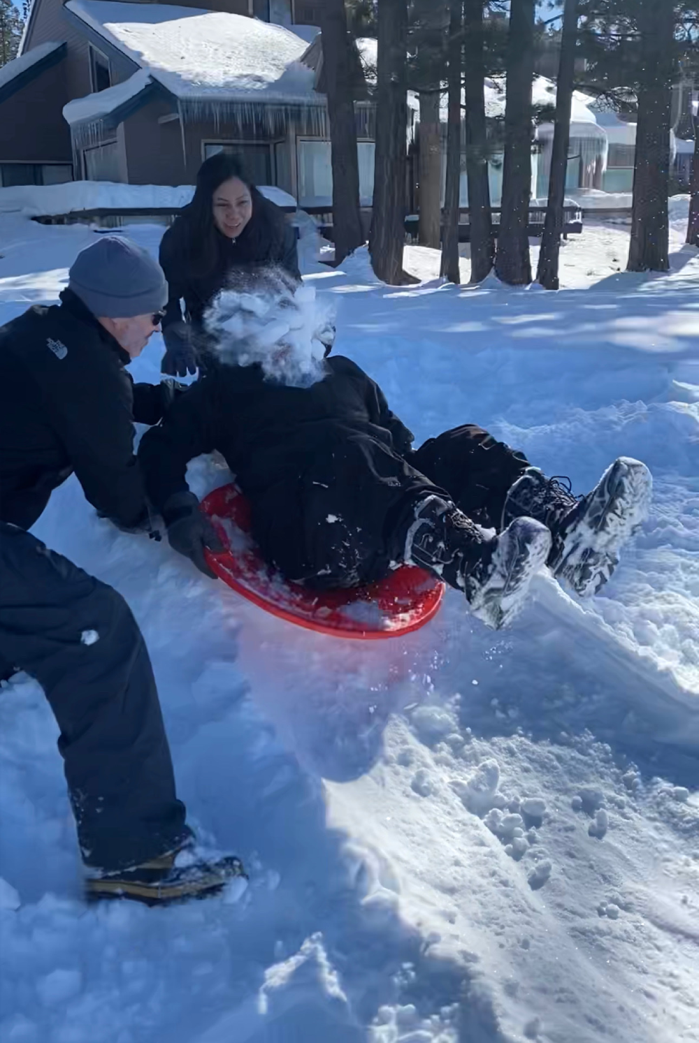 A person in a black coat sits on a red plastic sled while being pushed by two others, resulting in a face-full of snow.