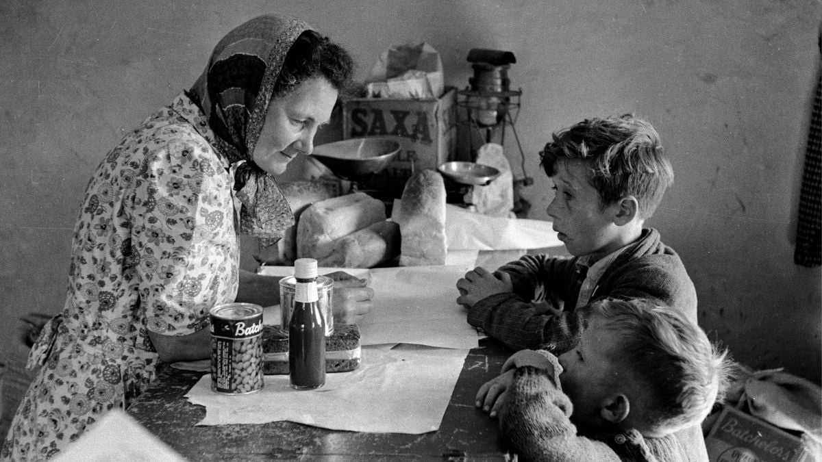 A woman in a headscarf sits at a table with two young boys, with groceries including Batchelors and Saxa branded items placed before them.