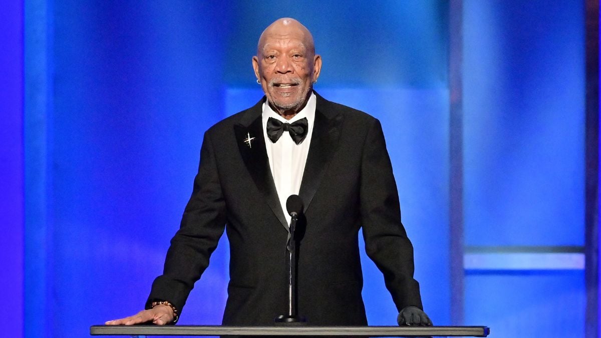 An older man in a black tuxedo and bow tie stands at a podium with a microphone, smiling, against a blue-lit background.