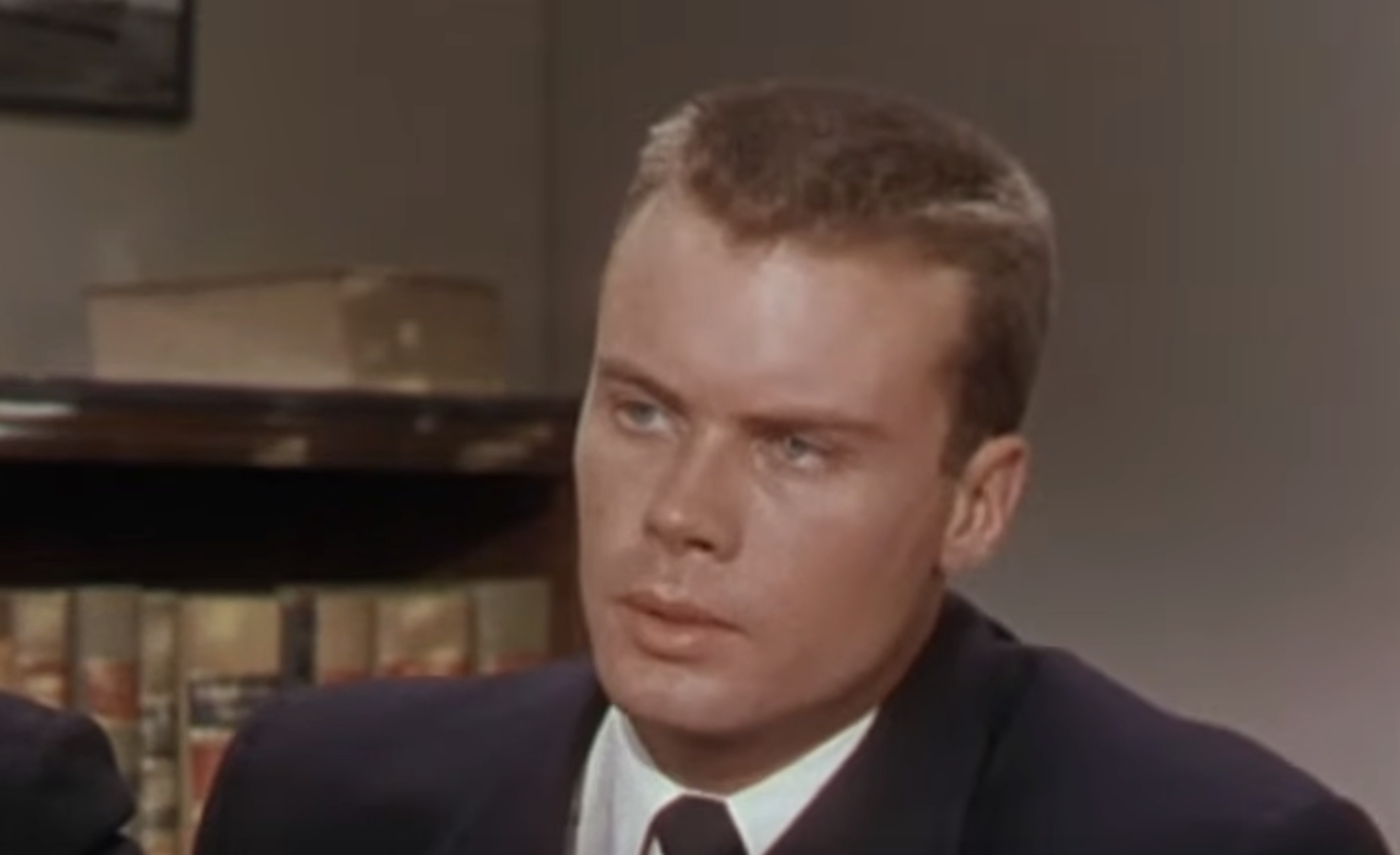 A man with short brown hair wearing a dark suit and tie looks forward with a serious expression.