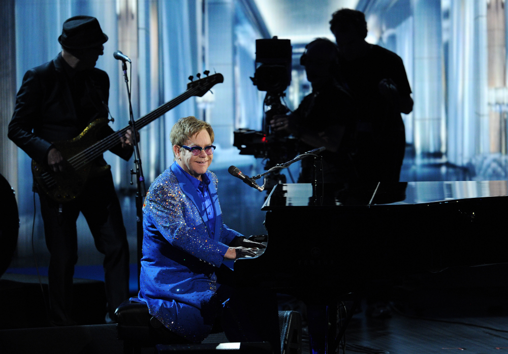Elton John performing at a piano wearing a blue sequined jacket, with a bassist and camera crew on stage behind him.