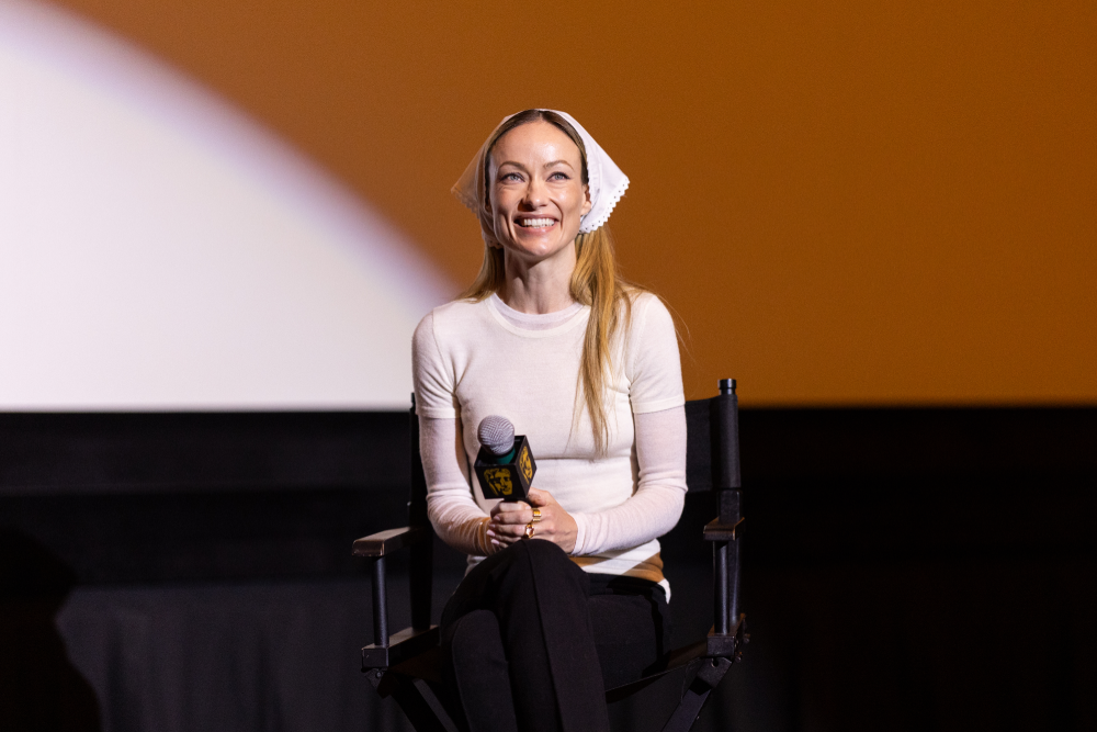 Olivia Wilde smiling while seated in a chair and holding a microphone on stage.