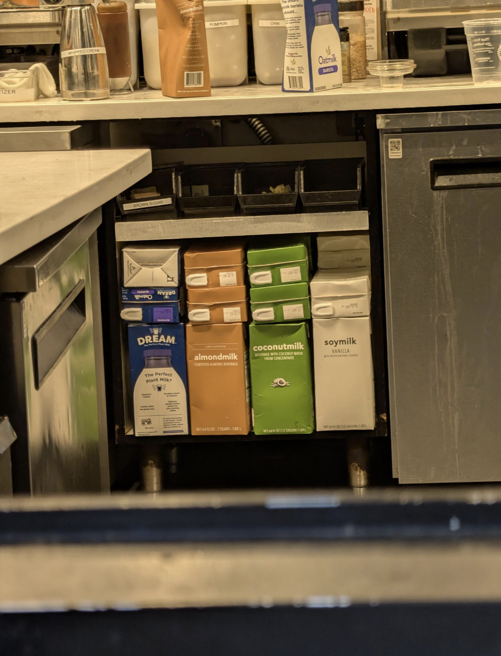 Cartons of almond milk, coconut milk, oat milk, soy milk, and rice milk are neatly arranged on a lower shelf under a counter in a café or kitchen, with other kitchen items visible above.