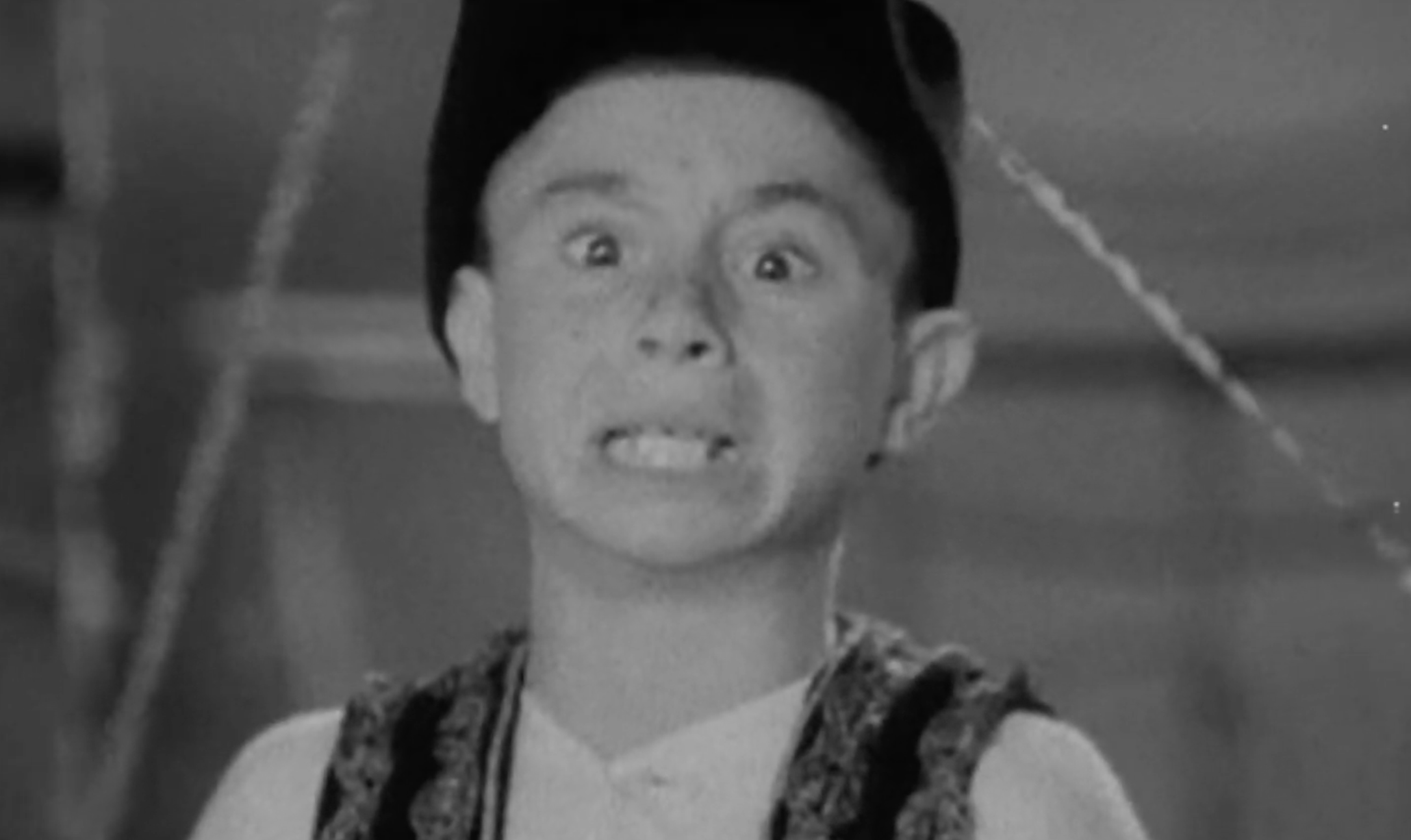 Black and white close-up of a young boy with a wide-eyed, frustrated expression wearing a small dark cap.