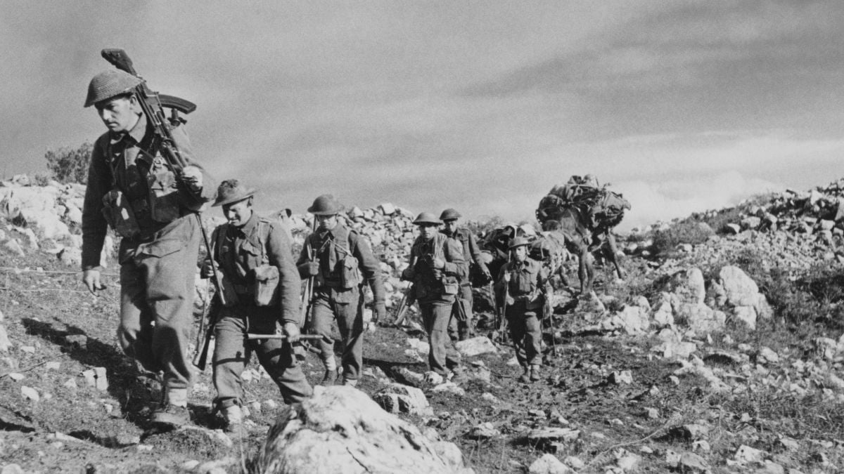 A group of soldiers in uniform and helmets walk in a rocky landscape, carrying gear and rifles. The sky is partly cloudy, and the soldiers appear focused and determined as they move uphill.