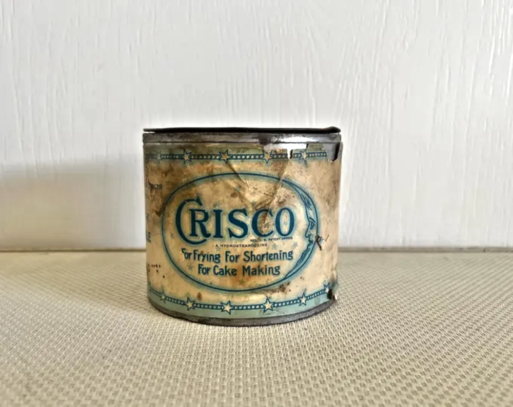A vintage, weathered Crisco shortening can with blue text reading “For Frying for Shortening for Cake Making.”