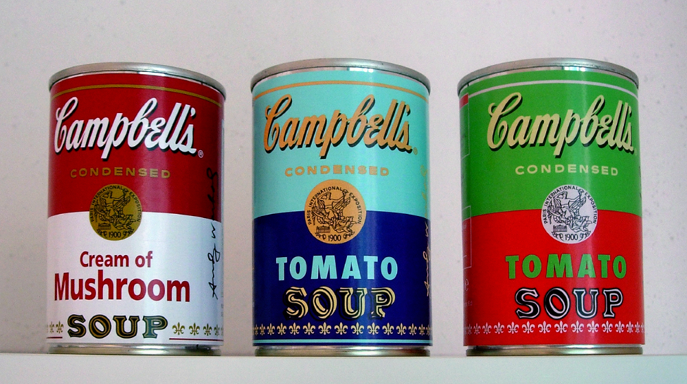 Three colorful Campbell’s condensed soup cans, labeled Cream of Mushroom, Tomato Soup, and Tomato Soup, sitting on a shelf.