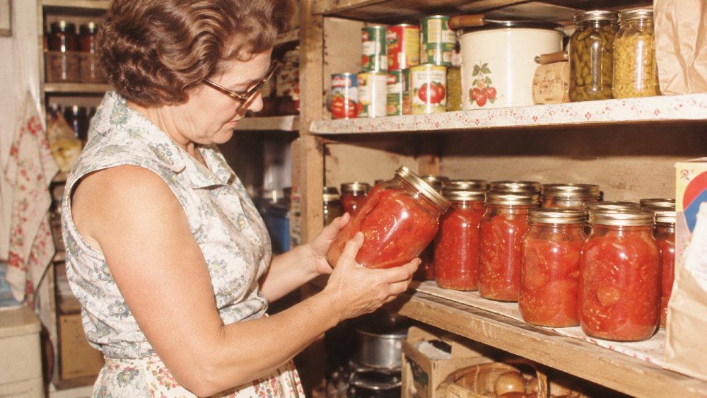 A woman in a floral dress examines a glass jar of home-canned tomatoes while standing in a pantry with shelves full of preserved food.