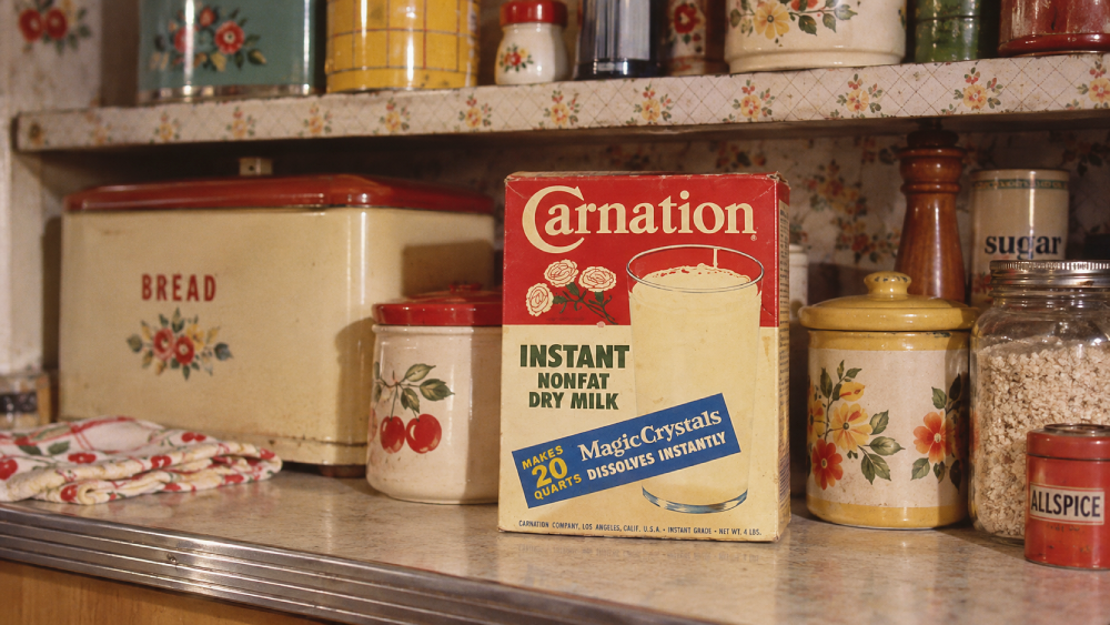 A vintage box of Carnation Instant Nonfat Dry Milk sits on a kitchen counter surrounded by floral canisters, a bread box, and spice jars.