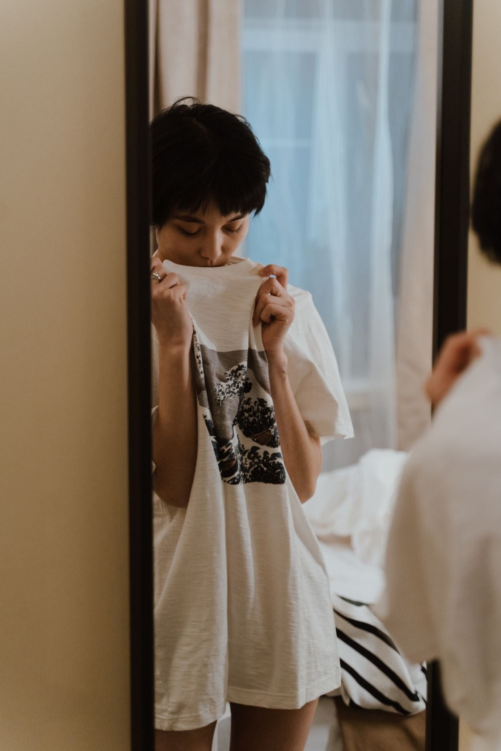 A person stands in front of a mirror, holding a white T-shirt with a black print close to their face, partially covering their mouth and nose, in a softly lit bedroom.
