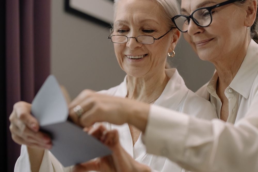Two older women in glasses and white shirts smile while looking at and holding open a greeting card together indoors.