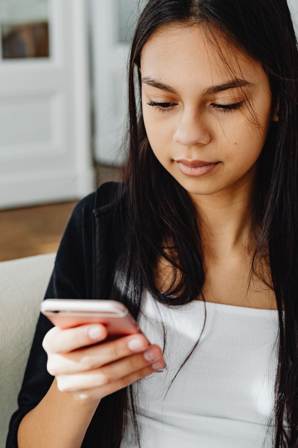A young woman with long dark hair looks down at her smartphone while sitting indoors, wearing a white top and black jacket.