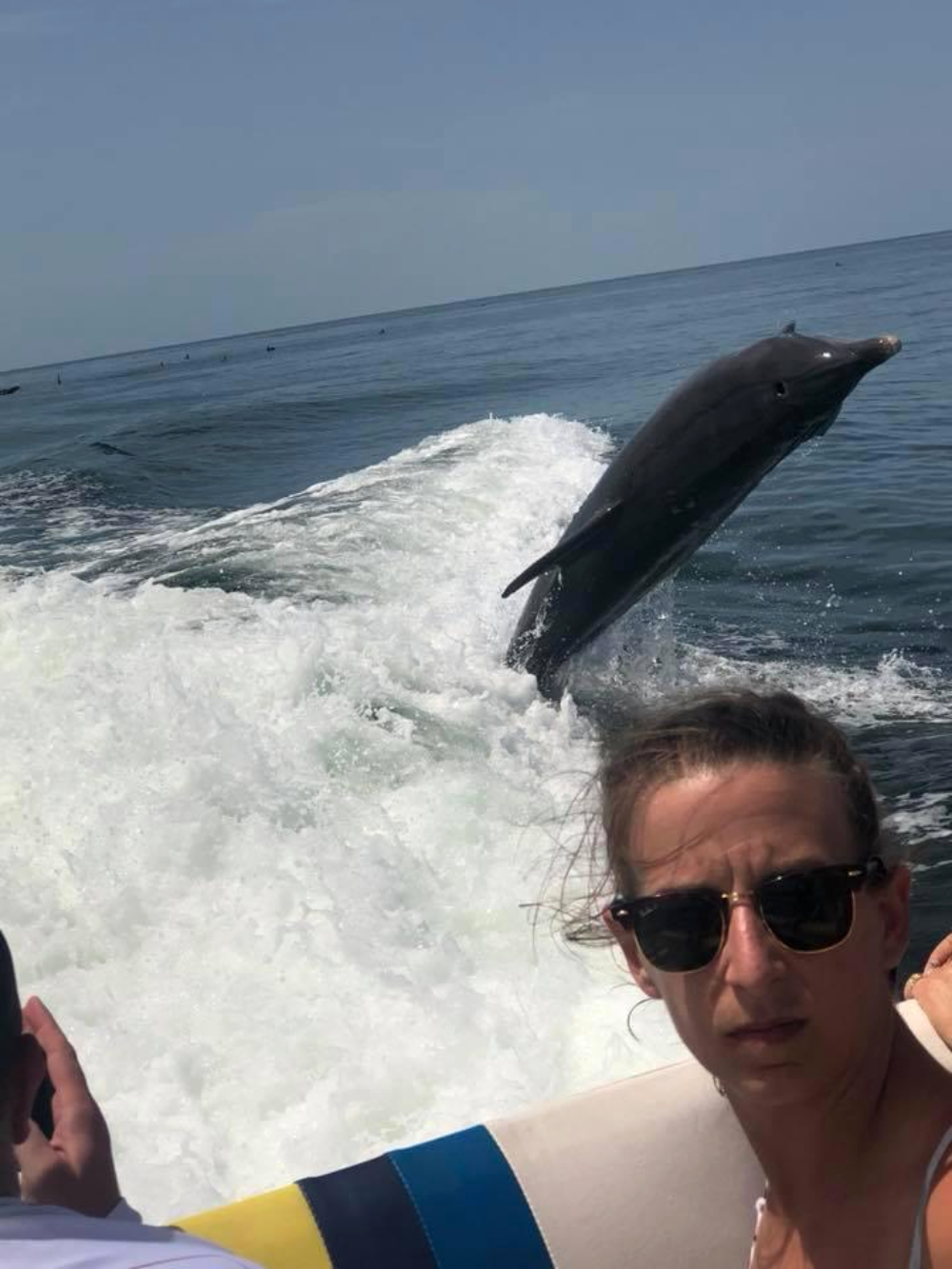 A woman wearing sunglasses takes a selfie on a boat as a dolphin leaps from the water behind her.