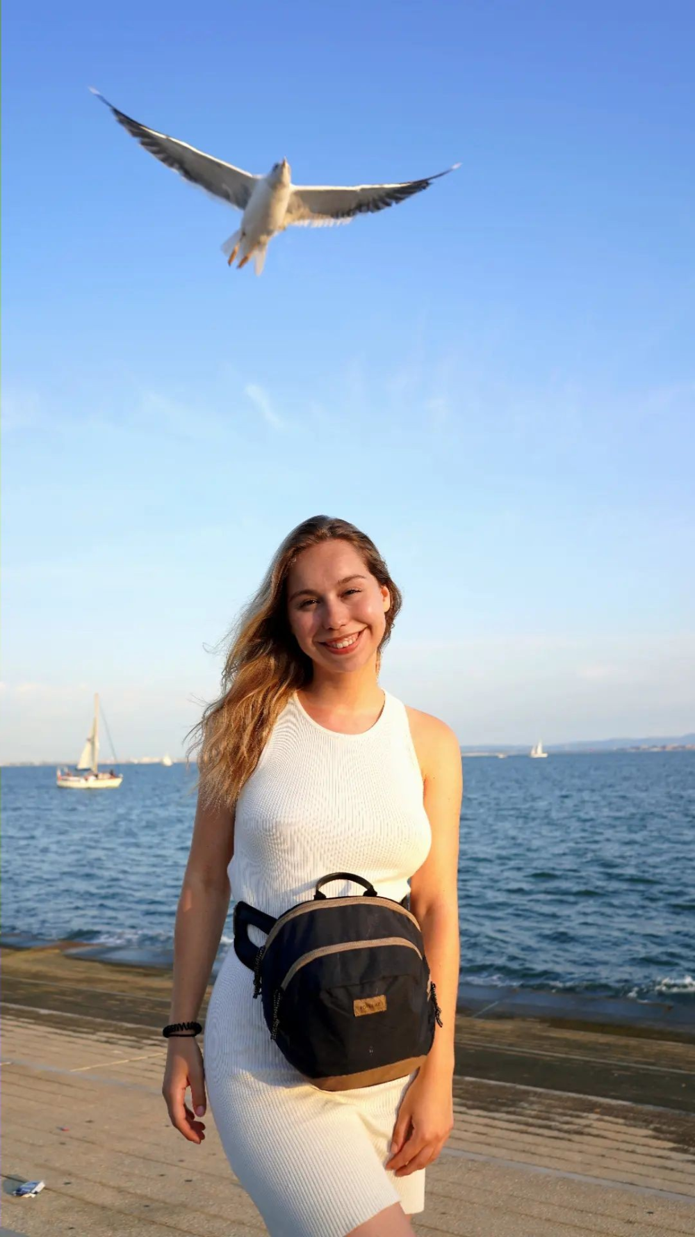 A smiling young woman in a white dress and small backpack stands on a waterfront promenade as a seagull flies overhead.